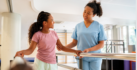 Physical therapist assists a child walking between parallel bars.
